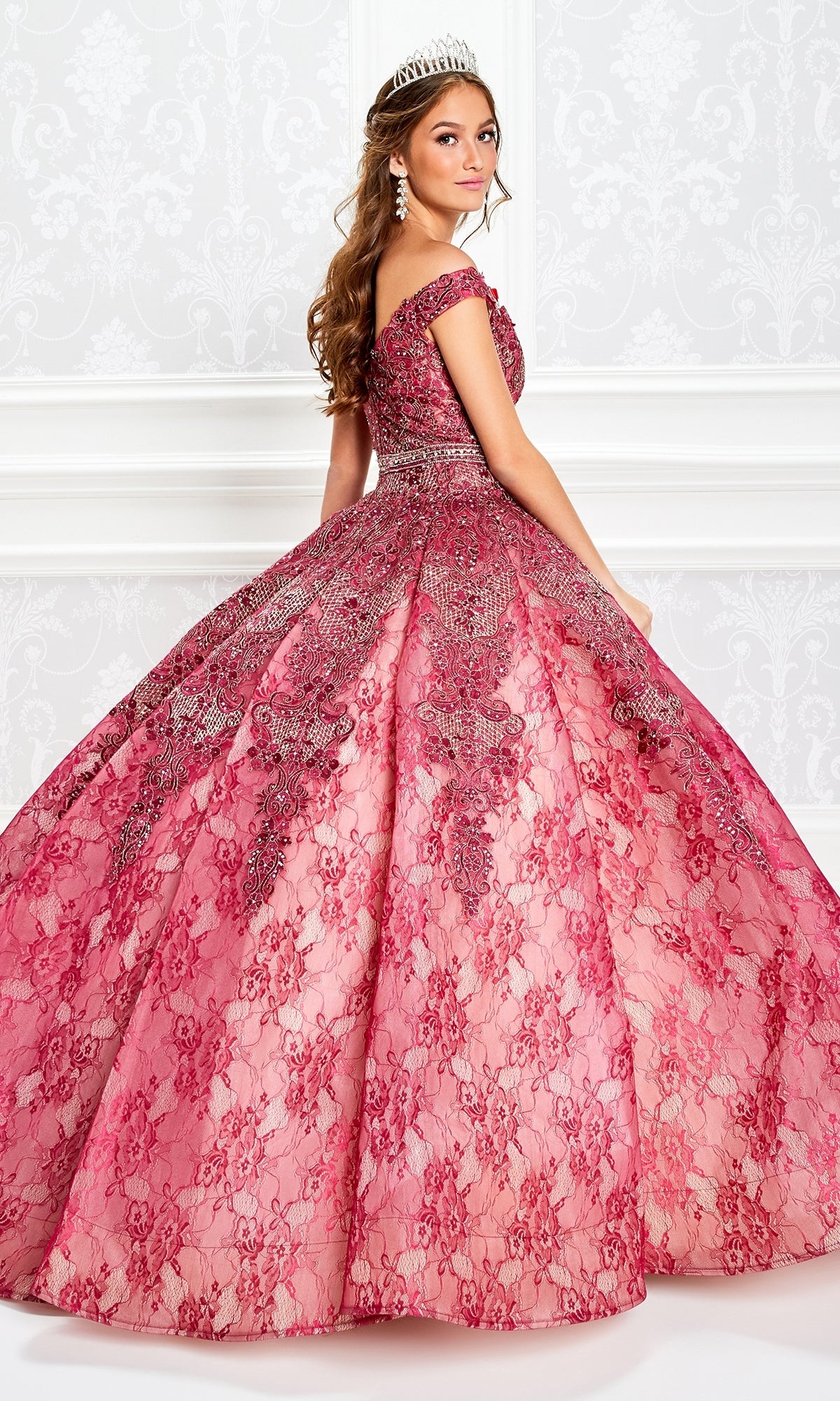 A young woman in a jeweled tiara wears the Princesa Lace Ball Gown Quinceañera Dress PR11921, featuring ornate beaded details and pink off-the-shoulder lace, posing against a white patterned wall.